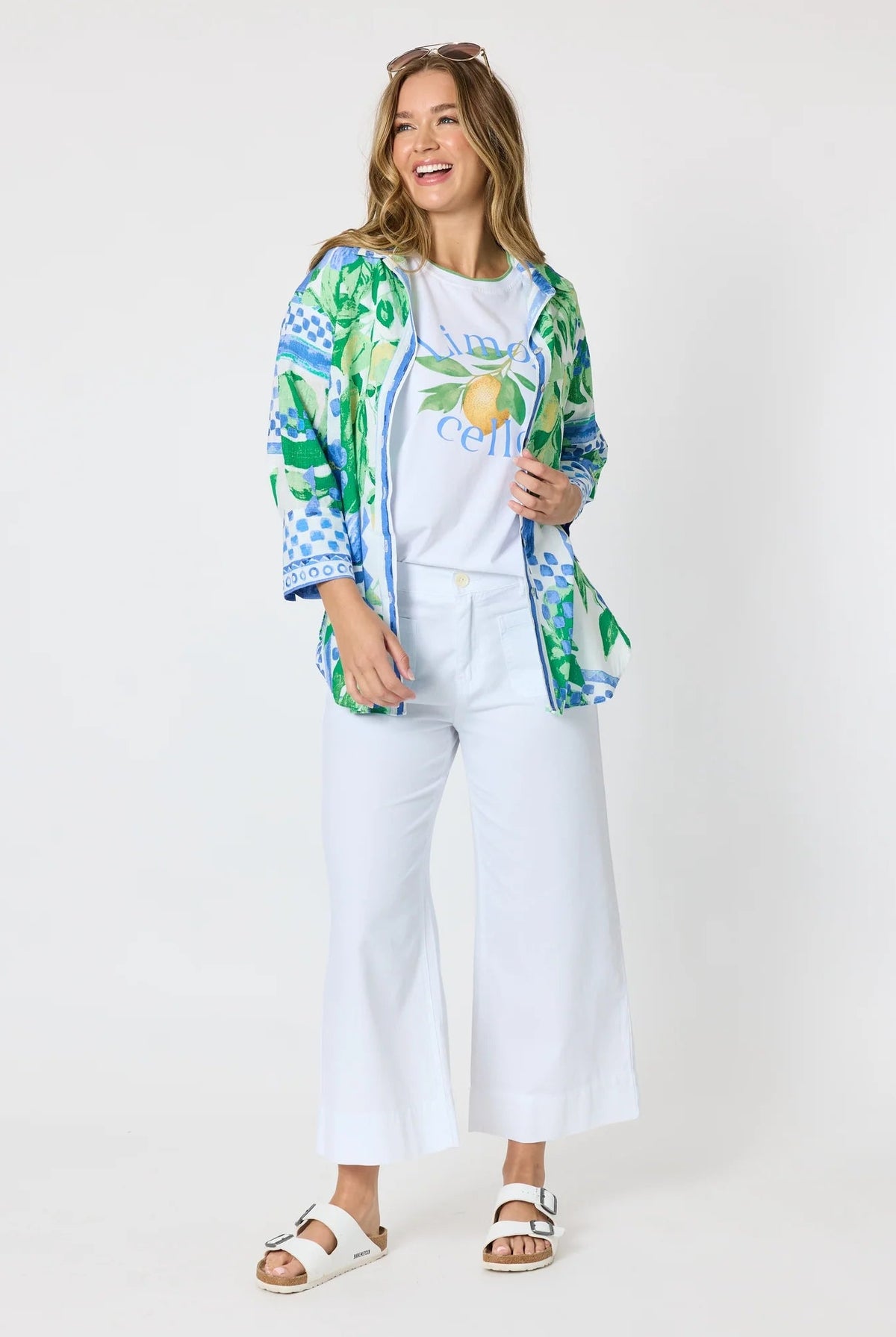 Woman wearing a button up shirt with collar and Mediterranean print with botanical, lemon and blue geometric design and white pants and white background.