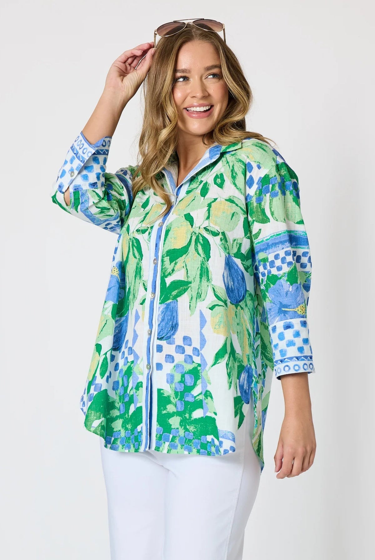 Woman wearing a button up shirt with collar and Mediterranean print with botanical, lemon and blue geometric design and white pants and white background.