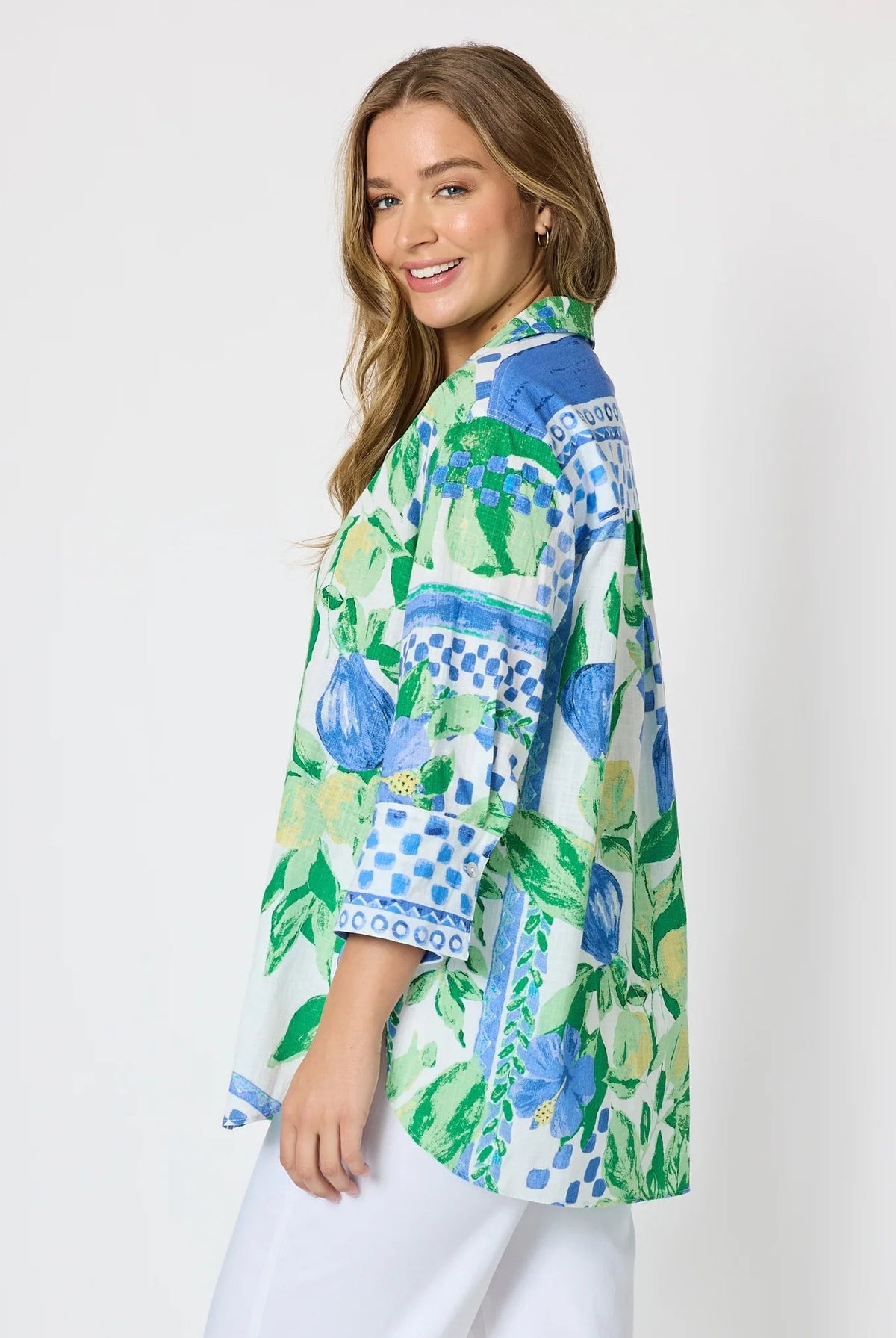 Woman wearing a button up shirt with collar and Mediterranean print with botanical, lemon and blue geometric design and white pants and white background.