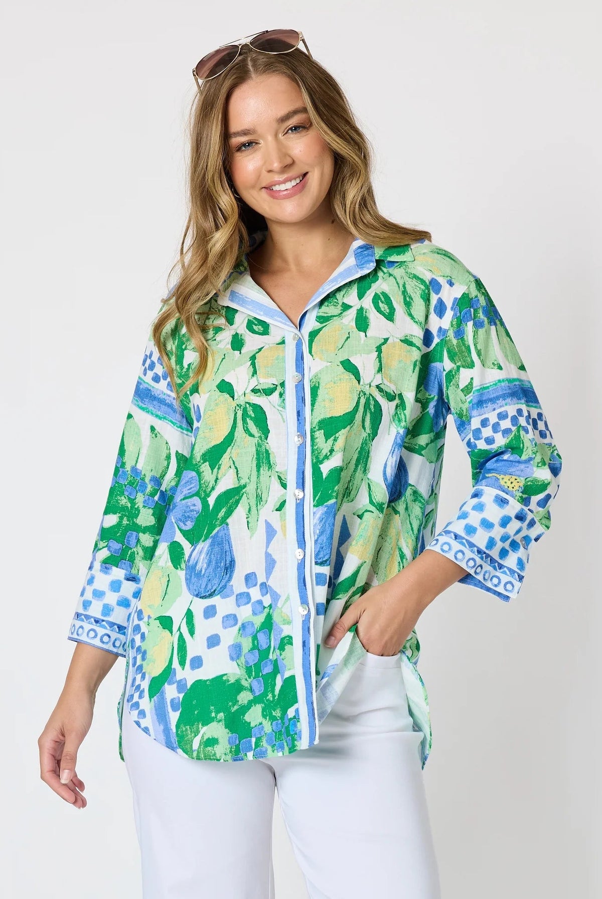 Woman wearing a button up shirt with collar and Mediterranean print with botanical, lemon and blue geometric design and white pants and white background.