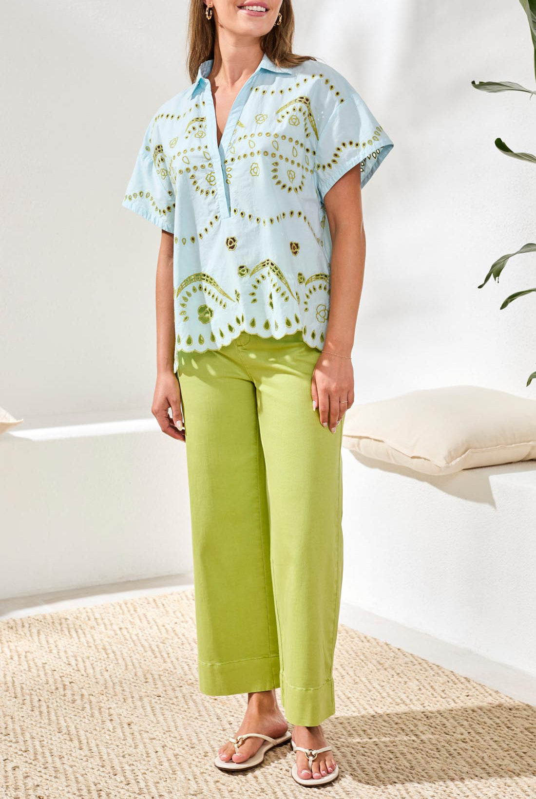 Woman wearing a light blue patterned top and green pants in a bright room.