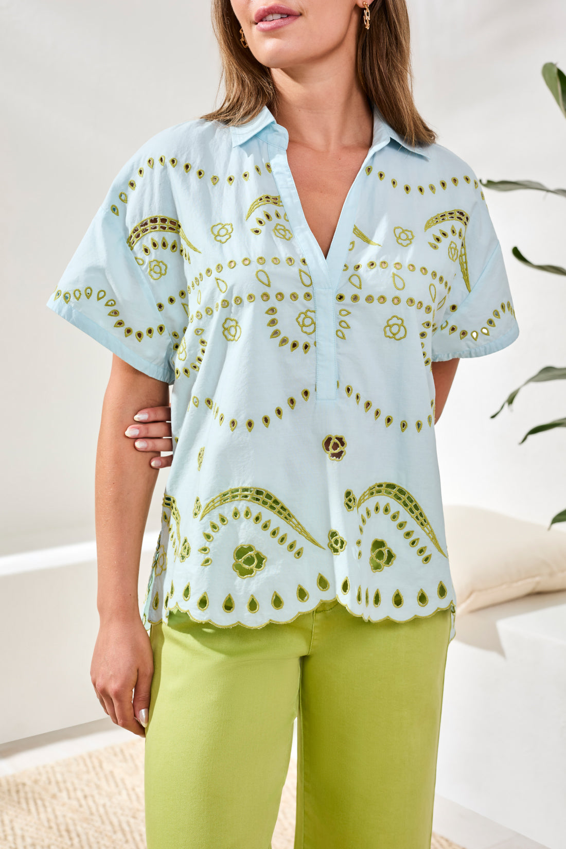Woman wearing a light blue shirt with green embroidery and green pants indoors.