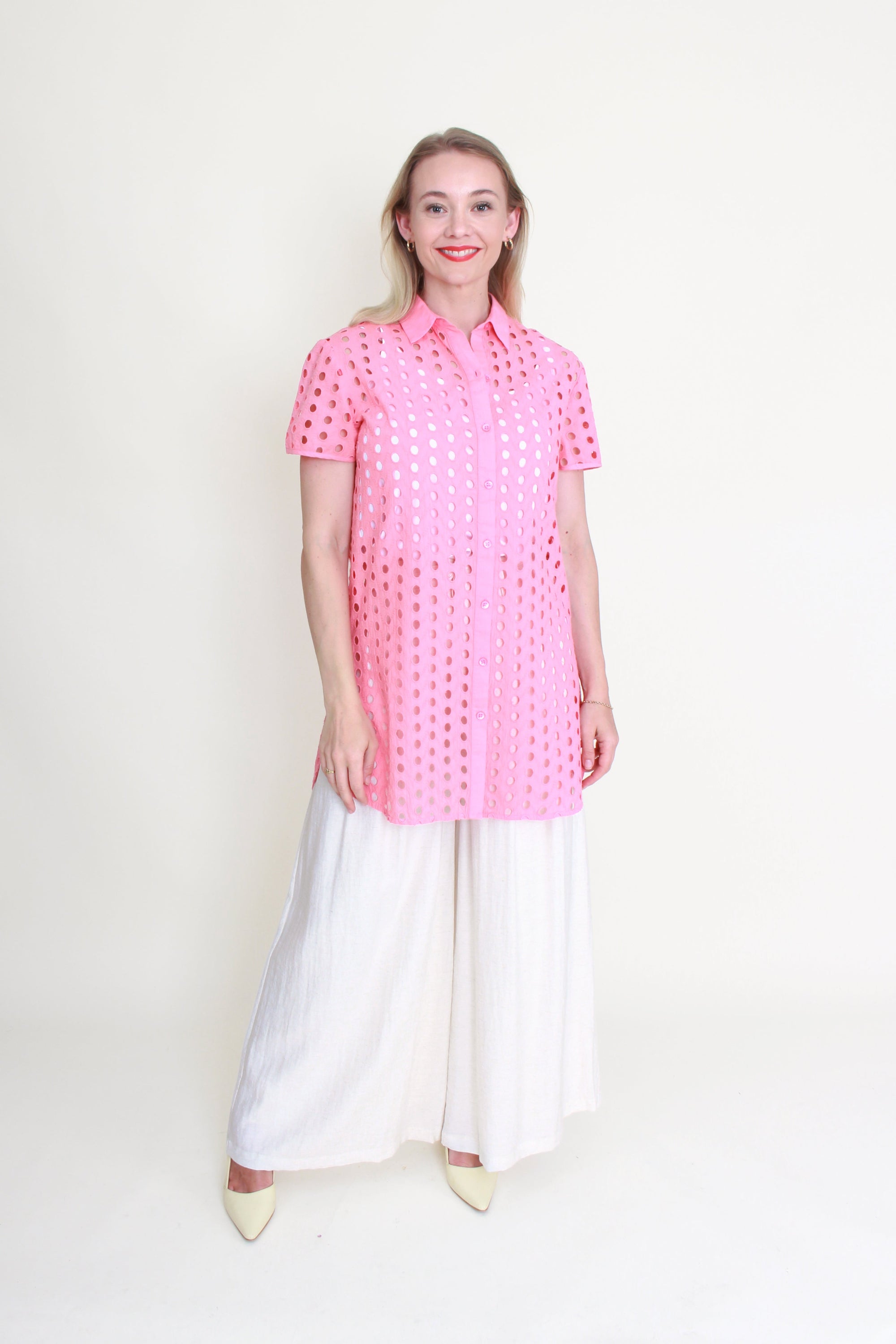 Woman wearing a pink anglaise shirt and white pants on a white background