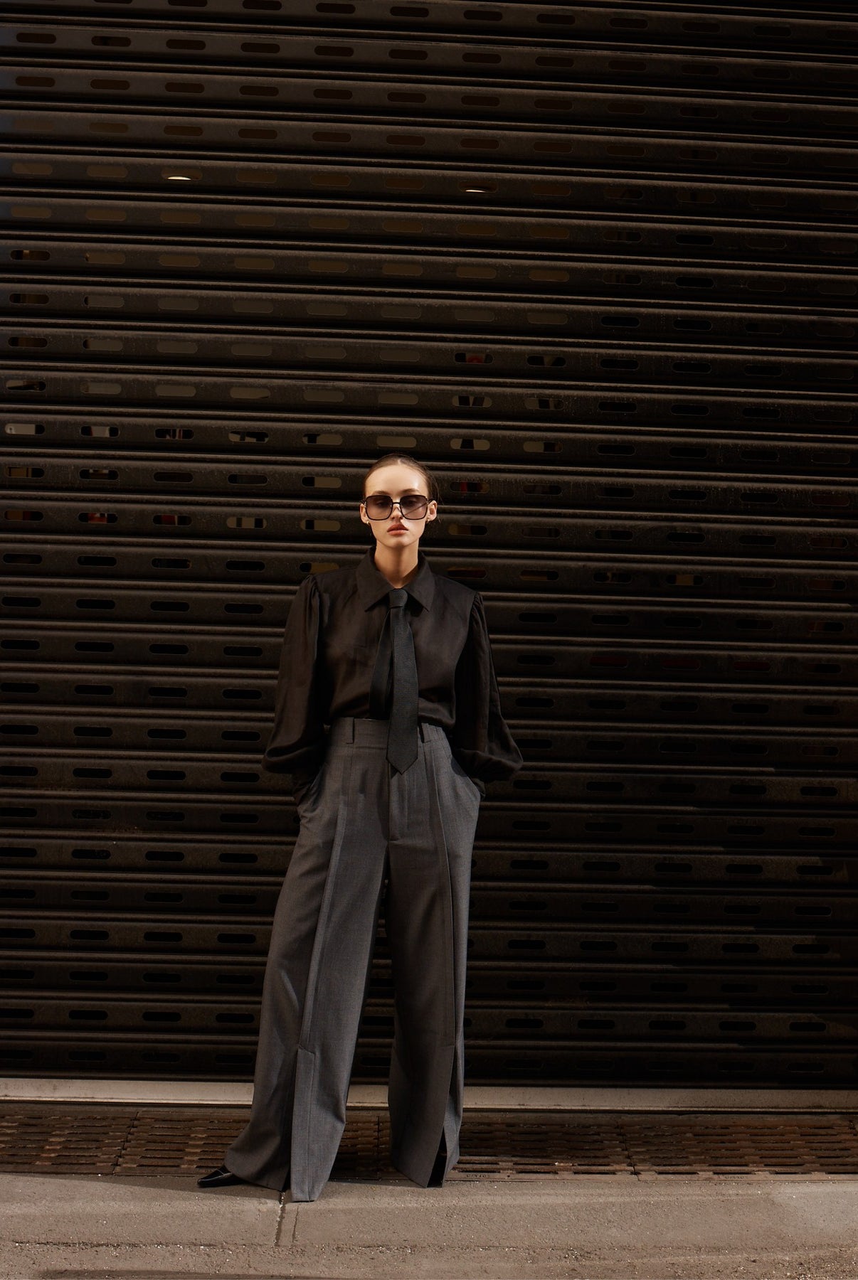 Person wearing a black shirt and gray pants standing in front of a dark metal shutter.