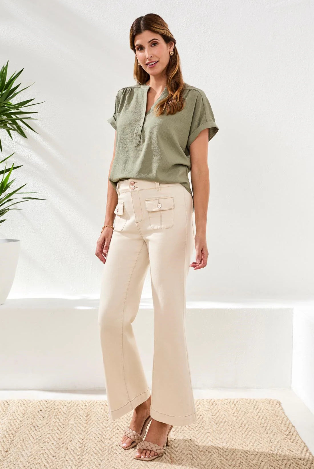Woman wearing a green top and beige pants standing in a bright room with plants.