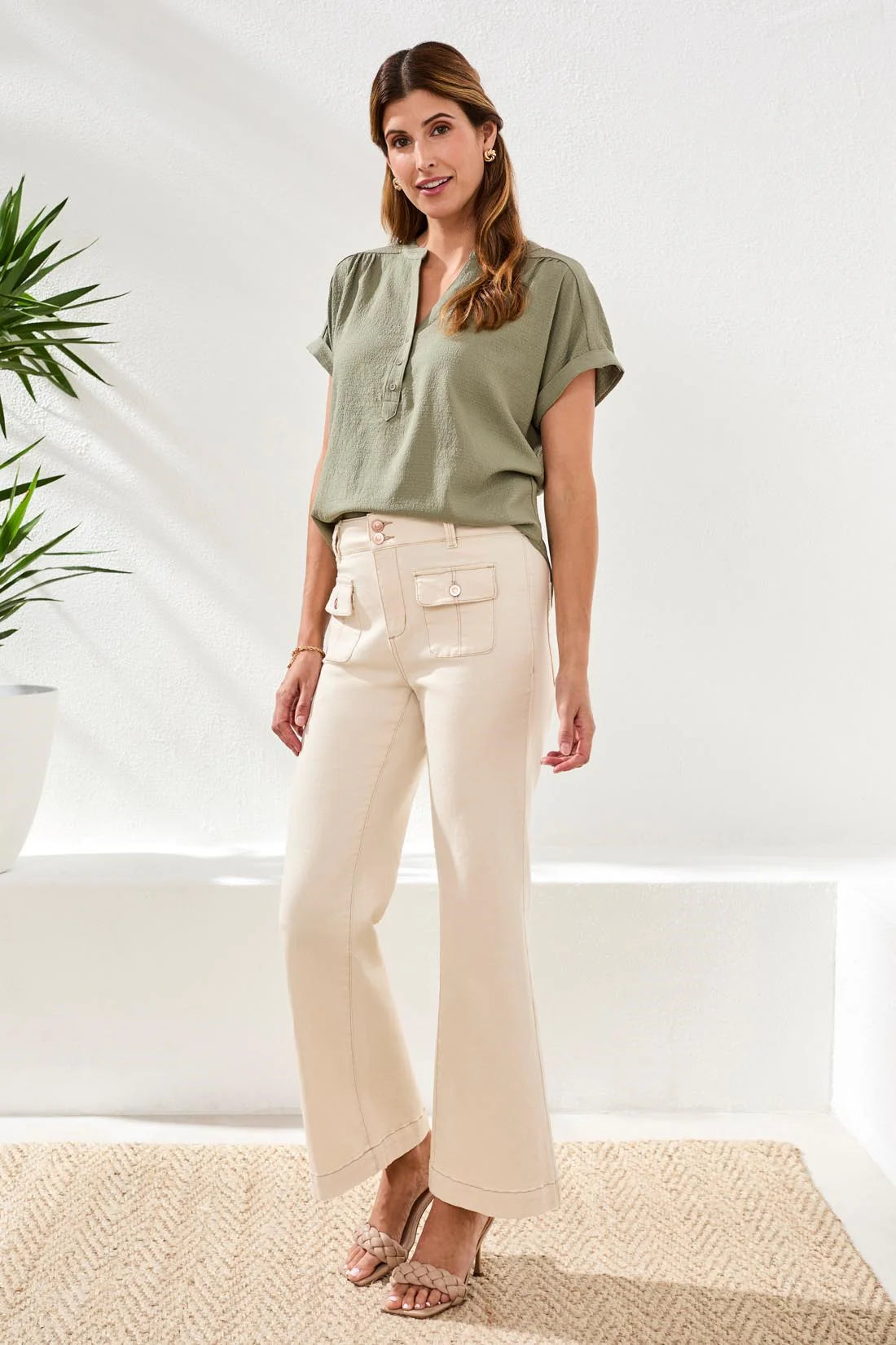 Woman wearing a green top and beige pants standing in a bright room with plants.
