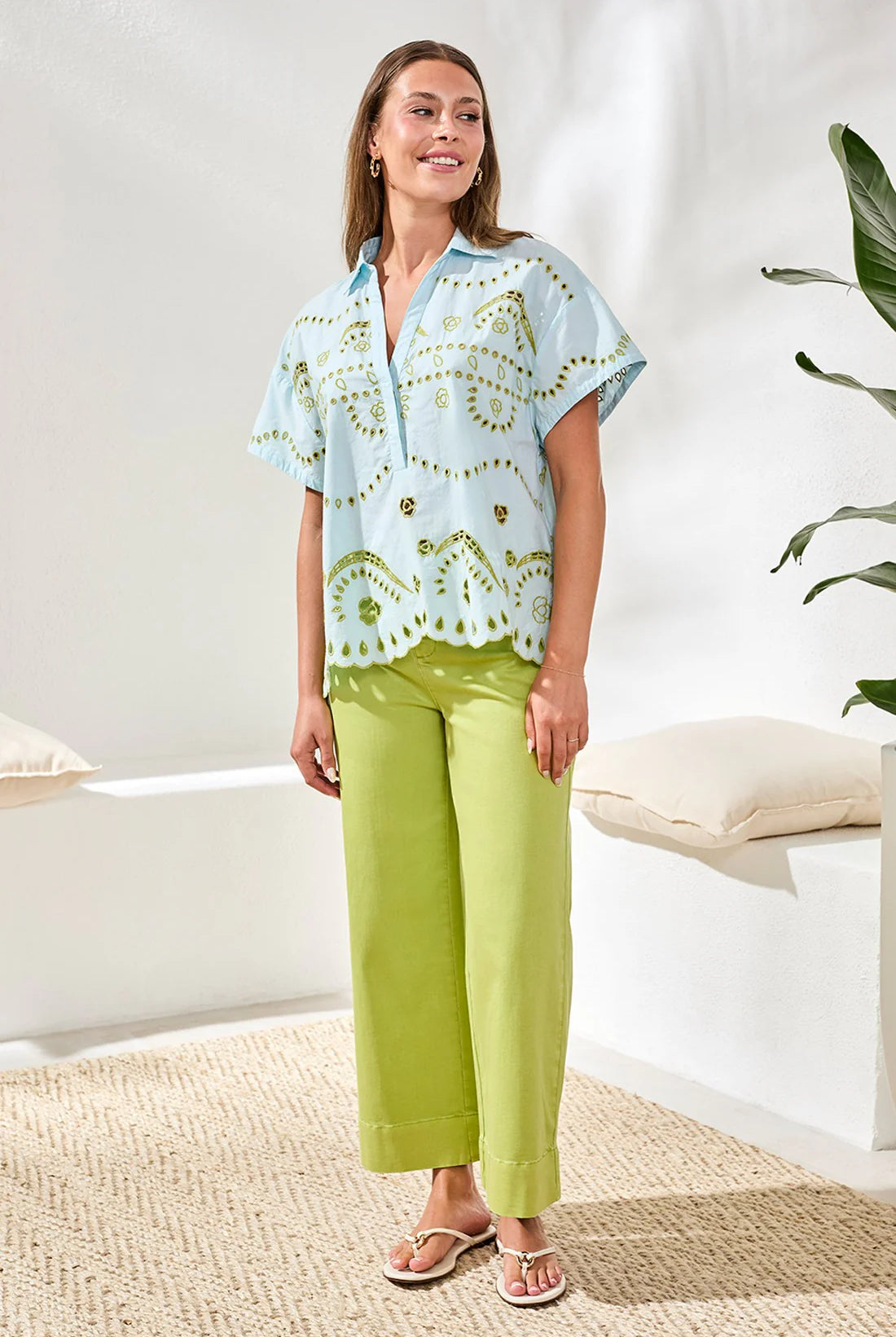Woman wearing a light blue patterned top and green pants in a bright room.