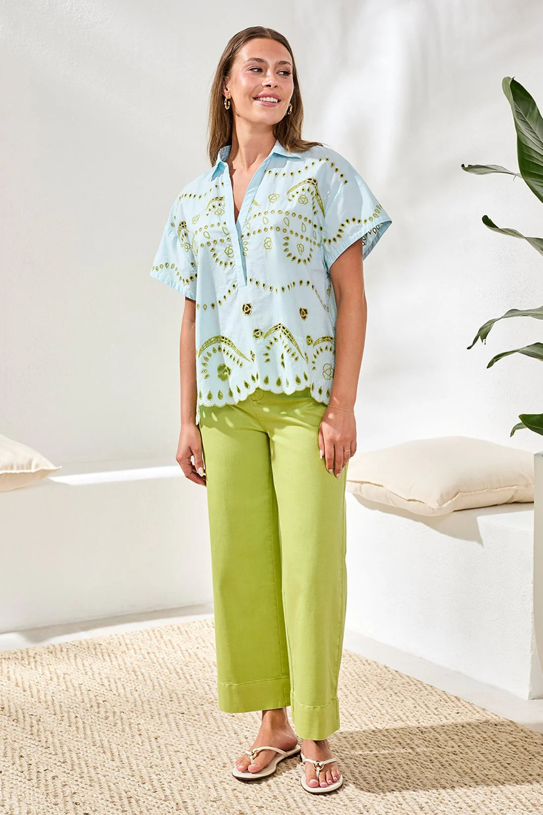 Woman wearing a light blue patterned top and green pants in a bright room.