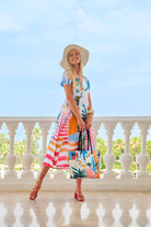 A woman stands on a sunlit terrace wearing a colorful tropical-print midi dress, a wide-brimmed straw hat, and carrying matching shopping bags. The background features a white balustrade railing and a clear blue sky.