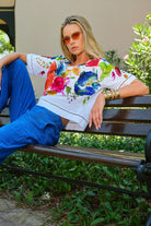 A woman wearing a white short-sleeve t-shirt with a vibrant multicolor floral print and bright blue pants, sitting on a wooden park bench.