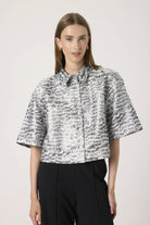 Woman wearing a patterned blouse and black pants against a light background