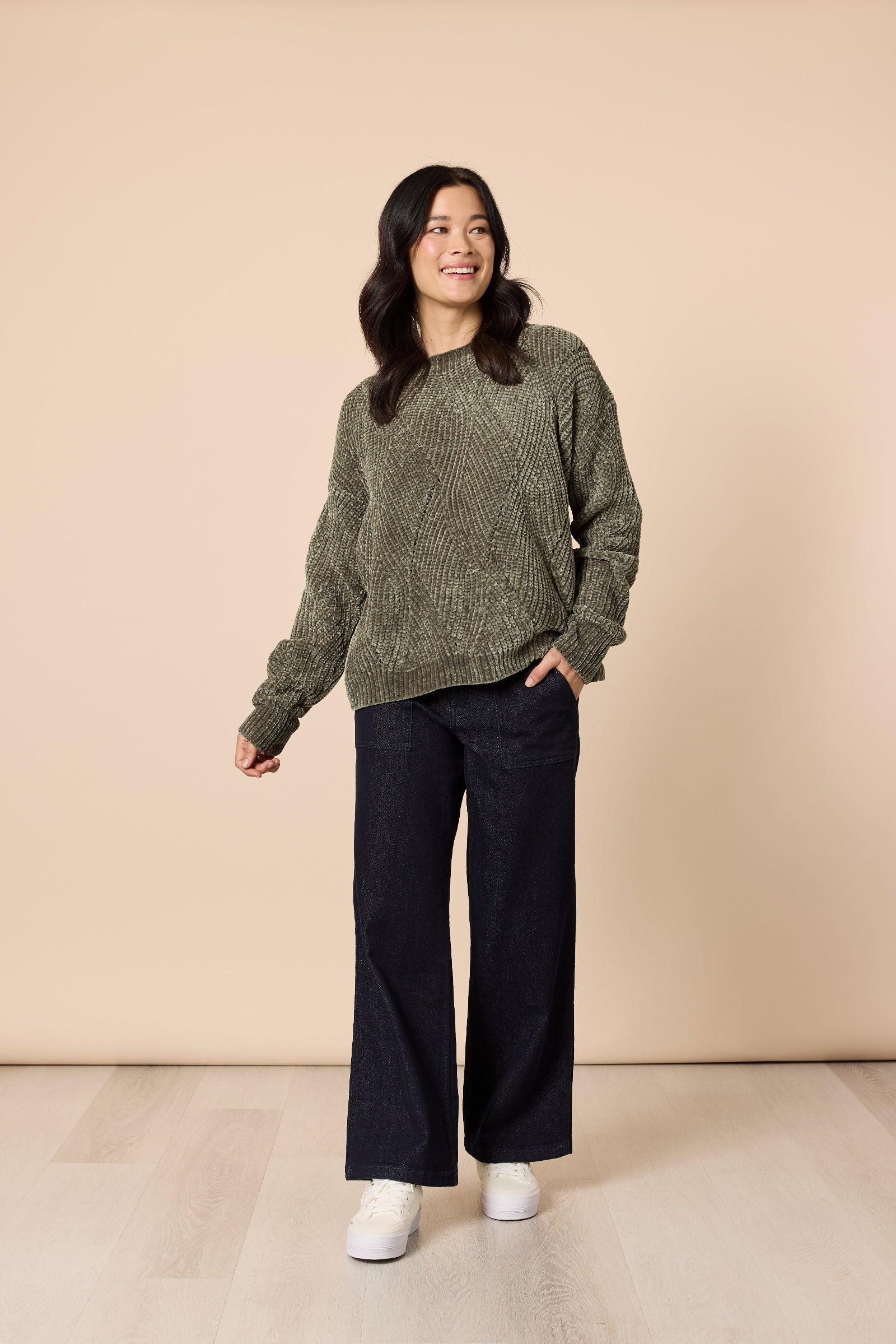 Woman wearing a green sweater and black pants against a beige background