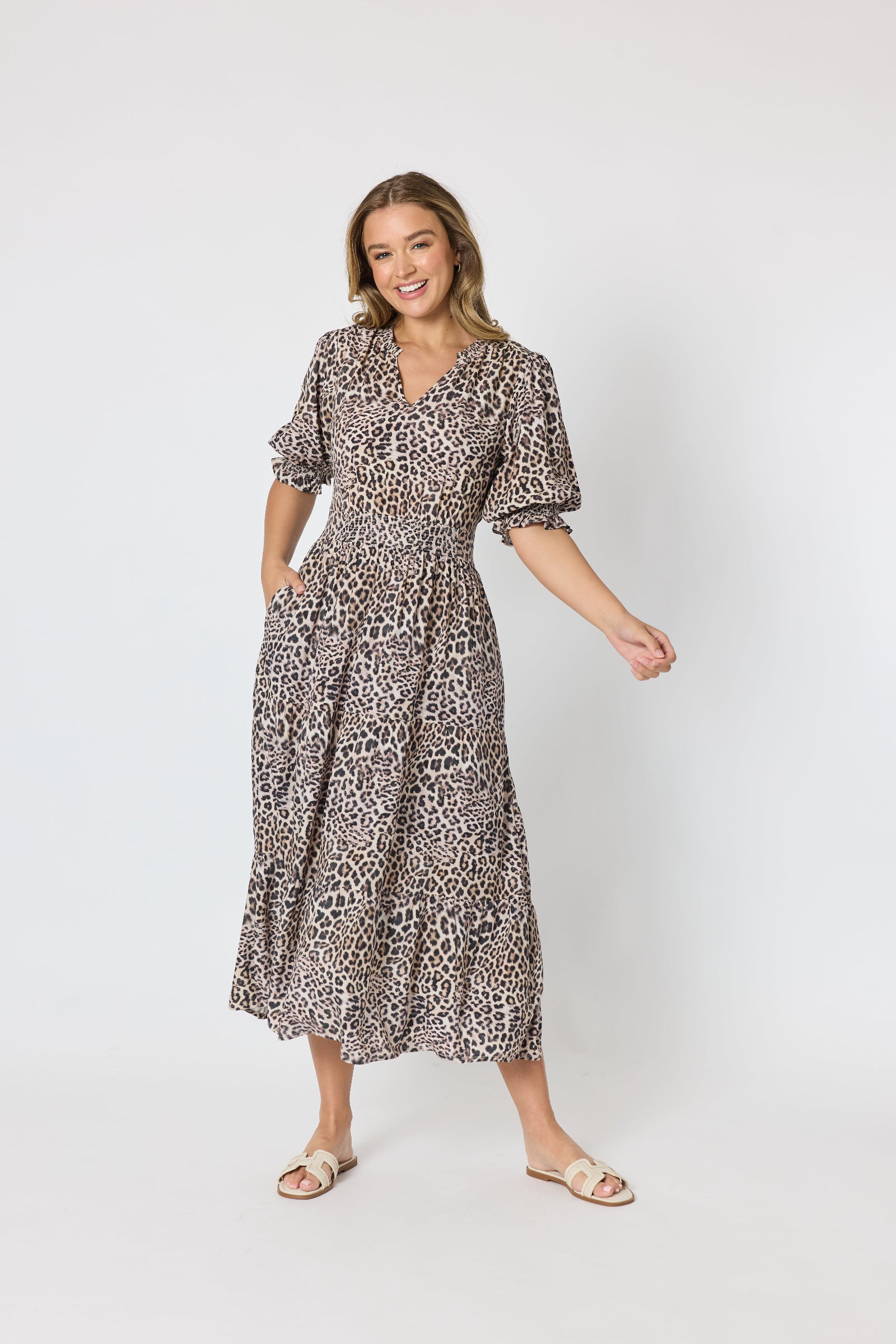 A woman modeling a leopard-print midi dress with puff sleeves and a V-neckline, paired with beige flat sandals, against a plain light gray studio background.