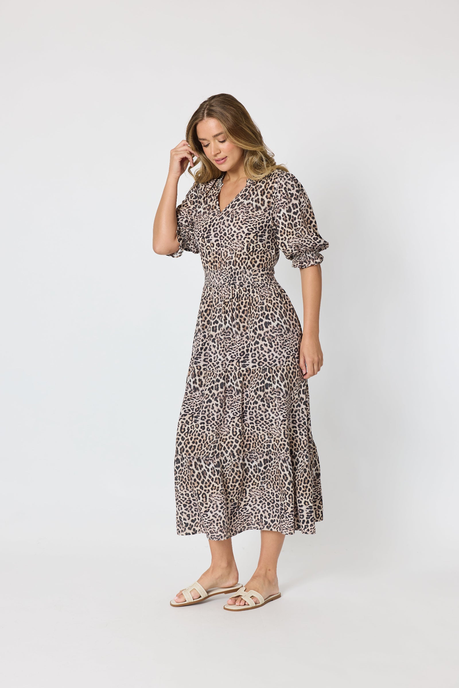 A woman modeling a leopard-print midi dress with puff sleeves and a V-neckline, paired with beige flat sandals, against a plain light gray studio background.