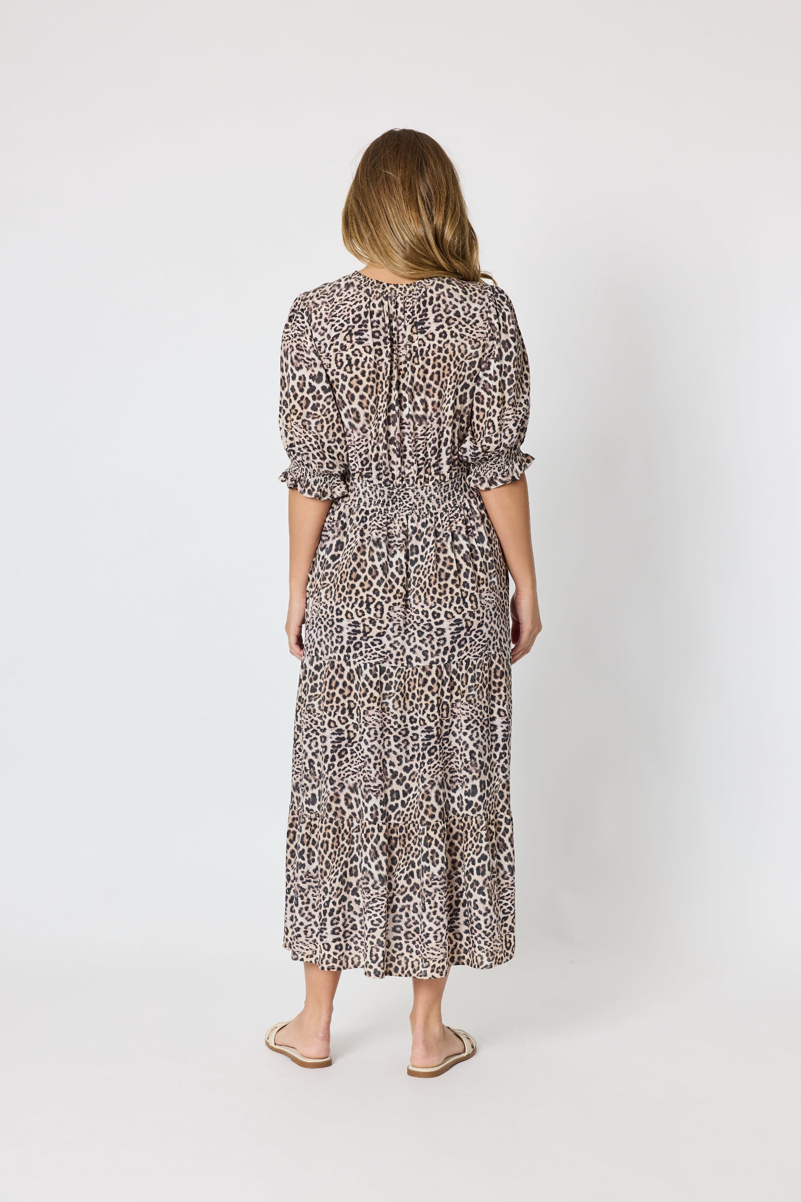 A woman viewed from behind wearing a leopard-print midi dress with puff sleeves and a tie at the back, paired with beige flat sandals, against a plain light gray studio background.