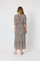 A woman viewed from behind wearing a leopard-print midi dress with puff sleeves and a tie at the back, paired with beige flat sandals, against a plain light gray studio background.