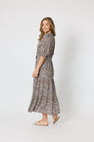 A woman modeling a leopard-print midi dress with three-quarter sleeves and a matching tie belt, standing against a plain light gray studio background.