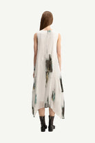 A woman viewed from behind wearing a sleeveless, high-low midi dress with an abstract watercolor print in green, black, and gray on a white base, paired with black ankle boots, against a solid off-white background.