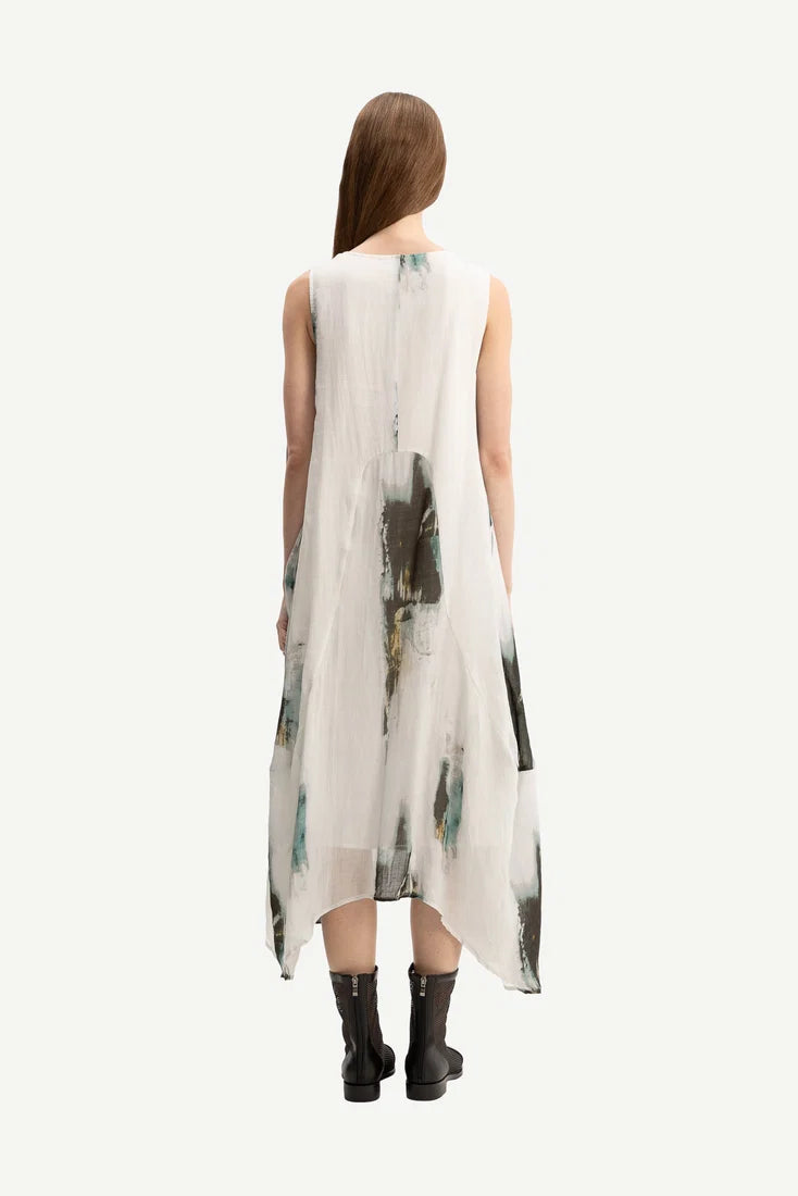 A woman viewed from behind wearing a sleeveless, high-low midi dress with an abstract watercolor print in green, black, and gray on a white base, paired with black ankle boots, against a solid off-white background.