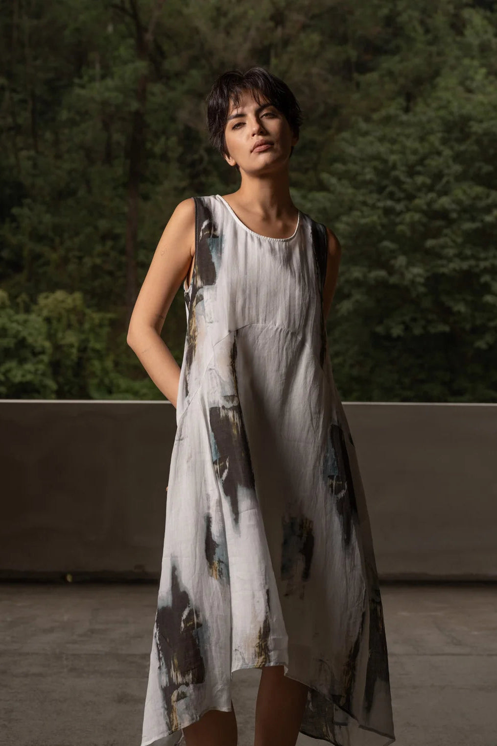 A woman wearing a sleeveless, high-low dress with an abstract painterly print in black, gray, and blue on a white base, standing outdoors against a backdrop of green trees and a concrete wall.