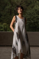 A woman wearing a sleeveless, high-low dress with an abstract painterly print in black, gray, and blue on a white base, standing outdoors against a backdrop of green trees and a concrete wall.