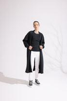 Person wearing a black long coat over a black sweater and white pants on a white background