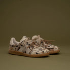 A pair of low-top sneakers with a beige, brown, and black spotted animal print upper, white laces, and gum soles, set against a solid olive green background.