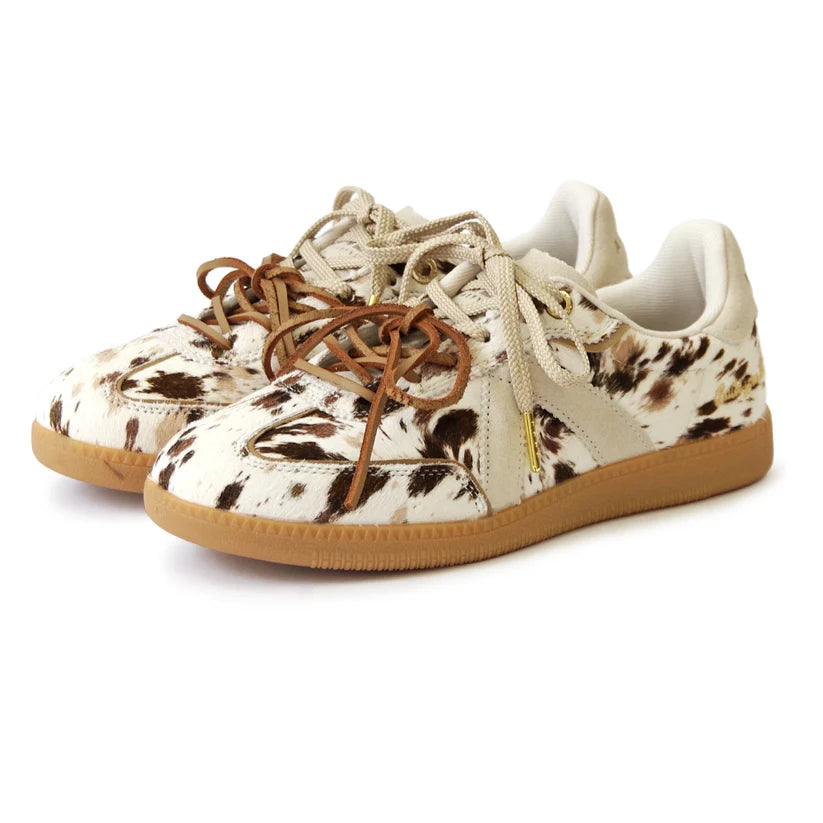 A pair of low-top sneakers featuring a cowhide-textured pattern in cream, brown, and black, with tan laces and gum soles, shown on a white background.