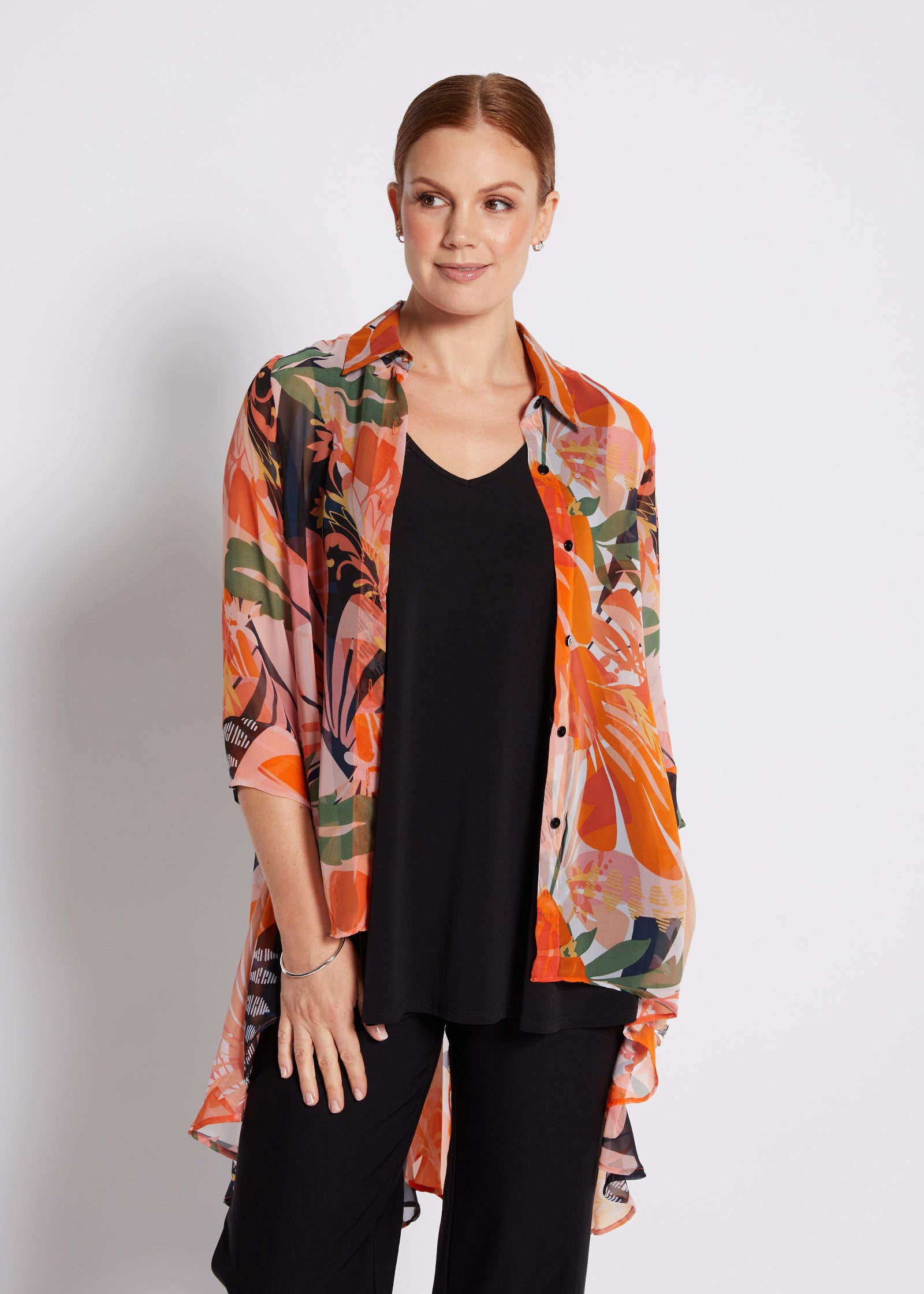 A woman modeling a lightweight, semi-sheer button-up shirt with a vibrant orange, pink, and green abstract floral print over a black top and black pants, set against a plain light gray studio background.