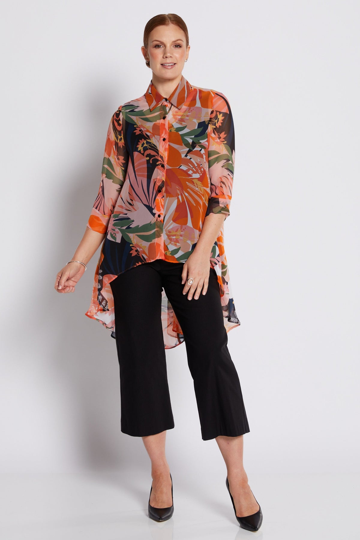 A woman modeling a long-sleeve button-up blouse with a vibrant orange, green, and black tropical floral print, paired with black cropped trousers and black heels, against a light gray studio background.