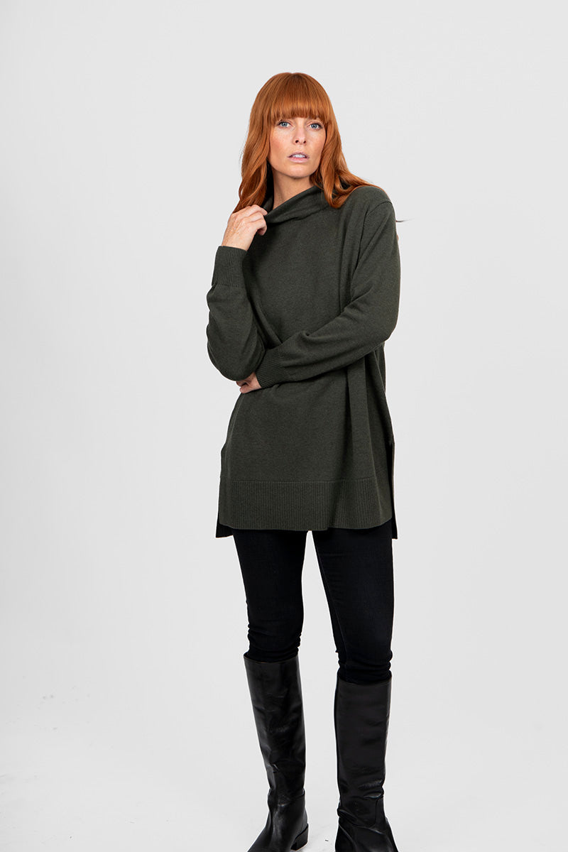 Woman wearing a long dark green sweater over a black top and pants on a white background