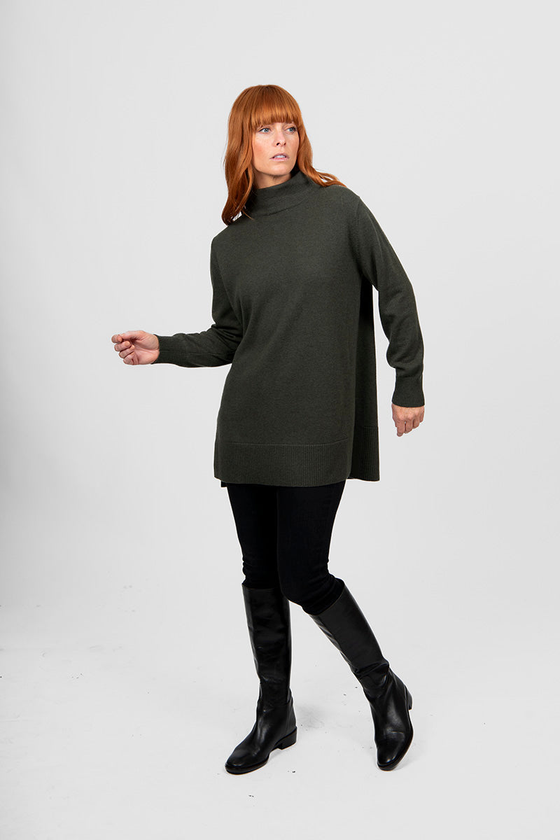 Woman wearing a long dark green turtleneck sweater and black pants on a white background