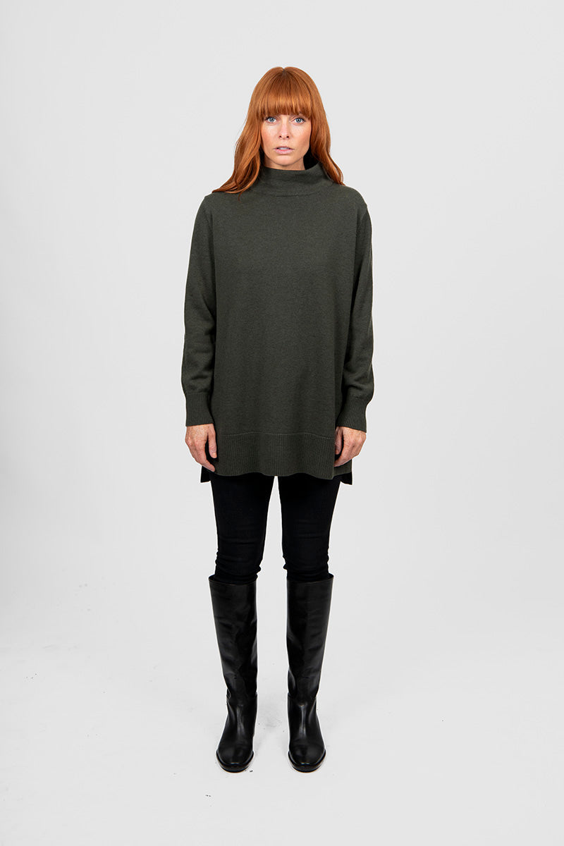 Woman wearing a long dark green turtleneck sweater and black pants on a white background