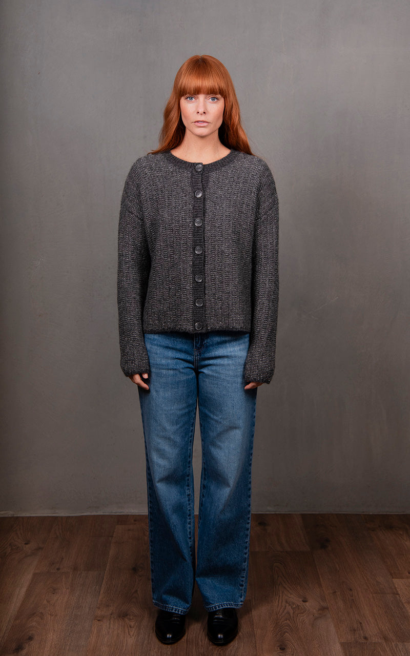 Woman wearing a gray textured cardigan, blue jeans, and black shoes against a gray background