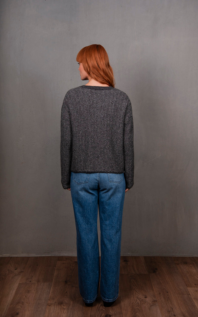 Person wearing a gray sweater and blue jeans standing against a plain gray wall.