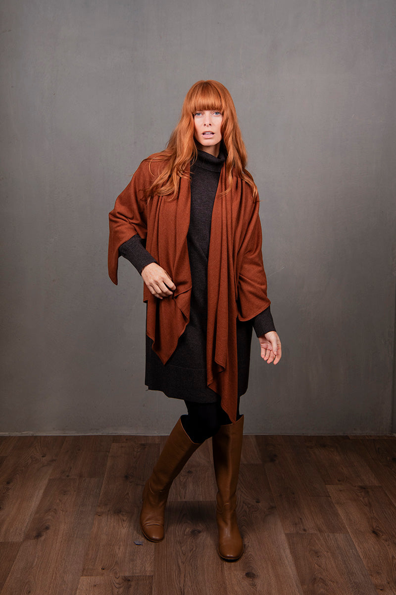 Woman wearing a brown poncho over a black outfit against a gray background