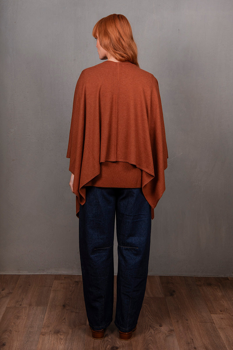 Person wearing a brown poncho and blue pants against a gray wall.