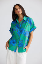 A woman wearing a vibrant green short-sleeve button-up shirt with a bold blue floral print, paired with white pants, photographed against a neutral light gray background.