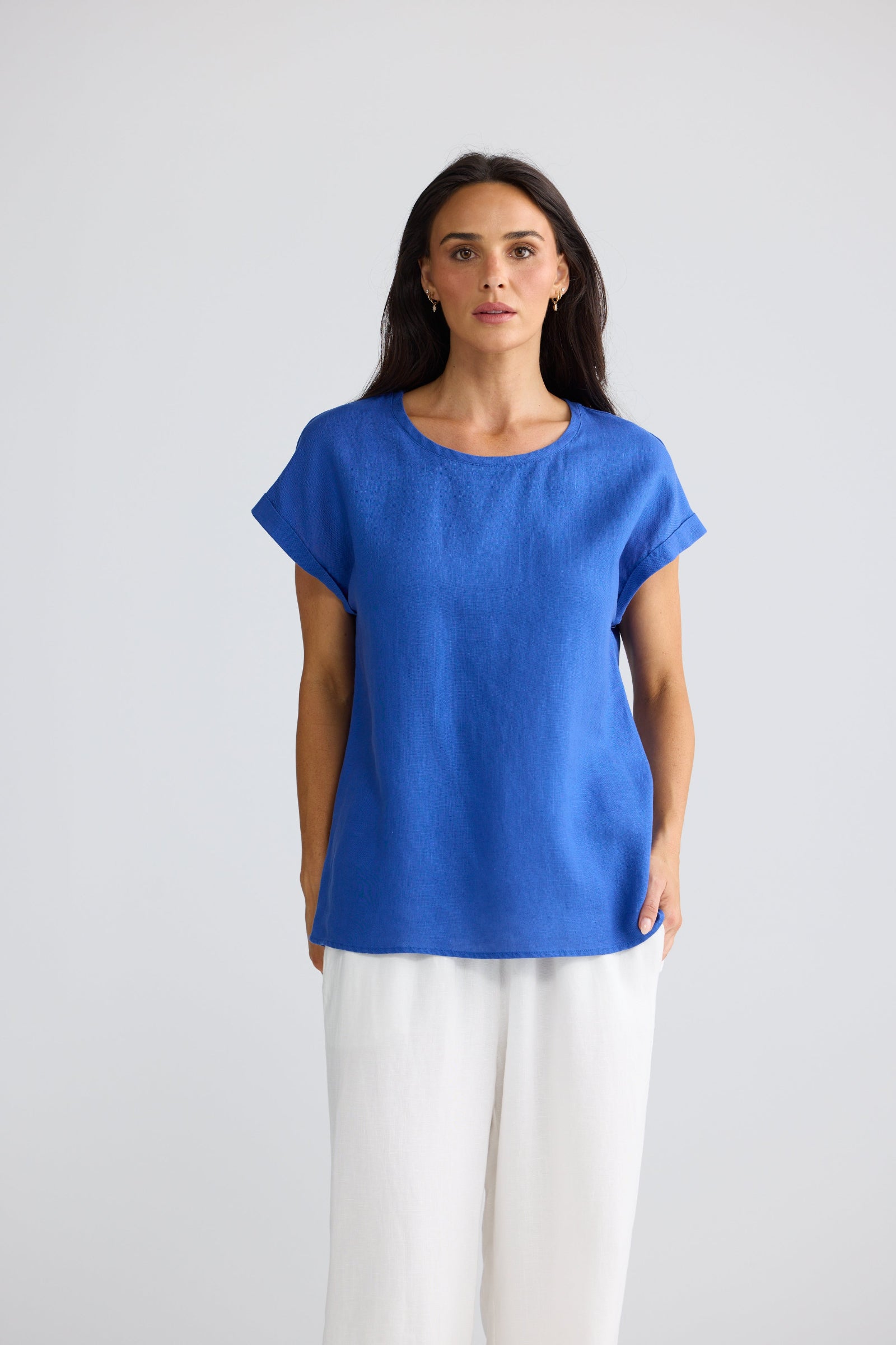 A woman modeling a bright blue short-sleeve top with a relaxed fit and white wide-leg pants, standing against a light gray studio background.