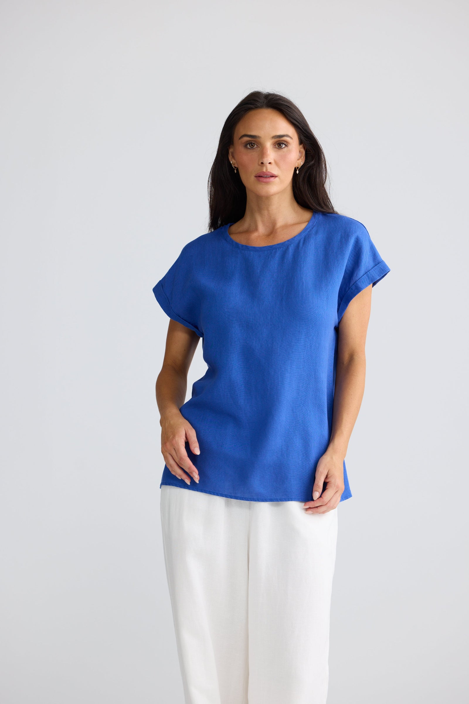 A woman modeling a short-sleeve, crew-neck top in a vibrant royal blue, paired with white wide-leg trousers, against a plain light gray studio background.