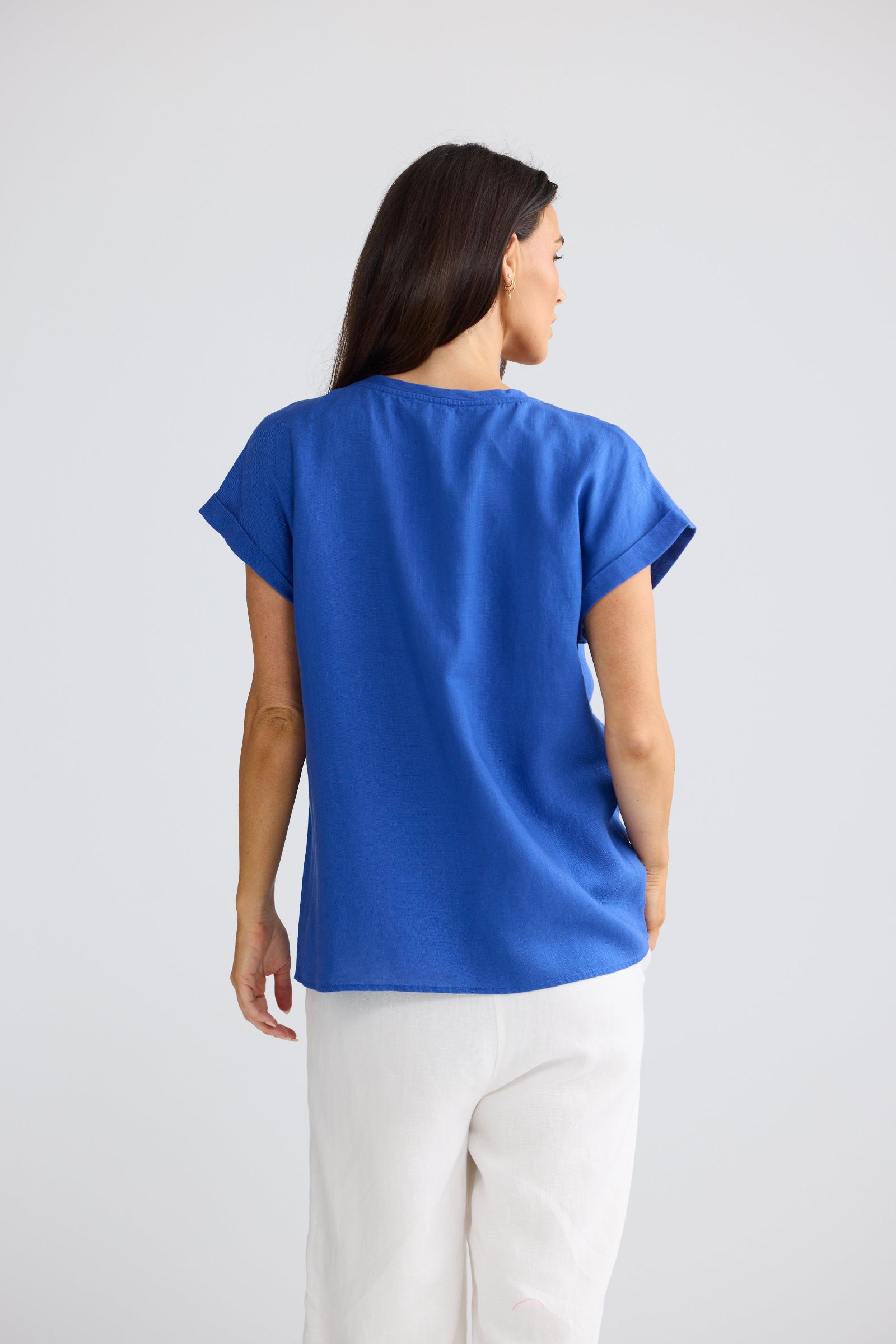 A woman shown from the back wearing a bright royal blue short-sleeve top and white pants against a light gray studio background.