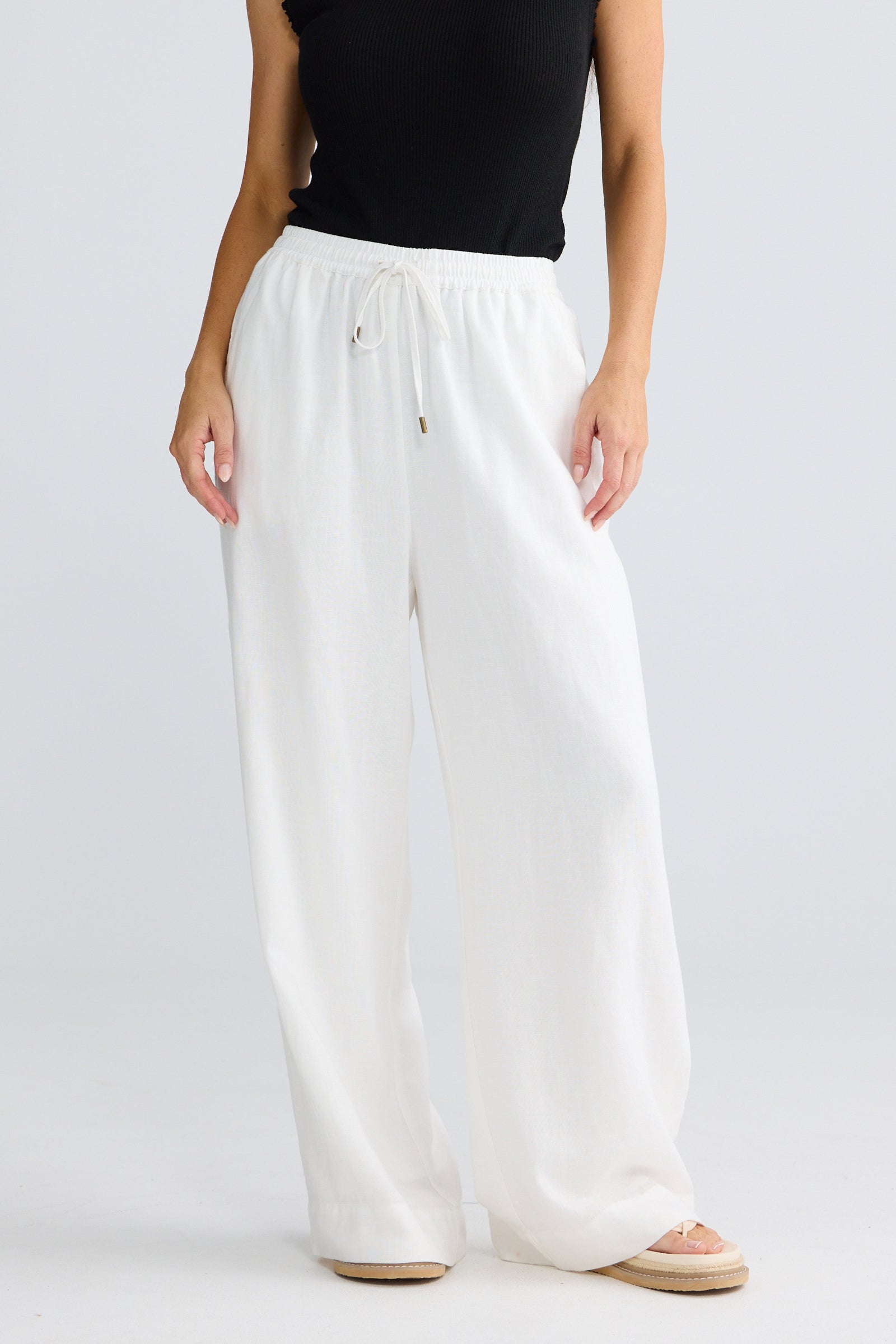 A model wearing white wide-leg drawstring pants with a black sleeveless top and beige flat sandals, shown from the chest down against a light gray studio background.