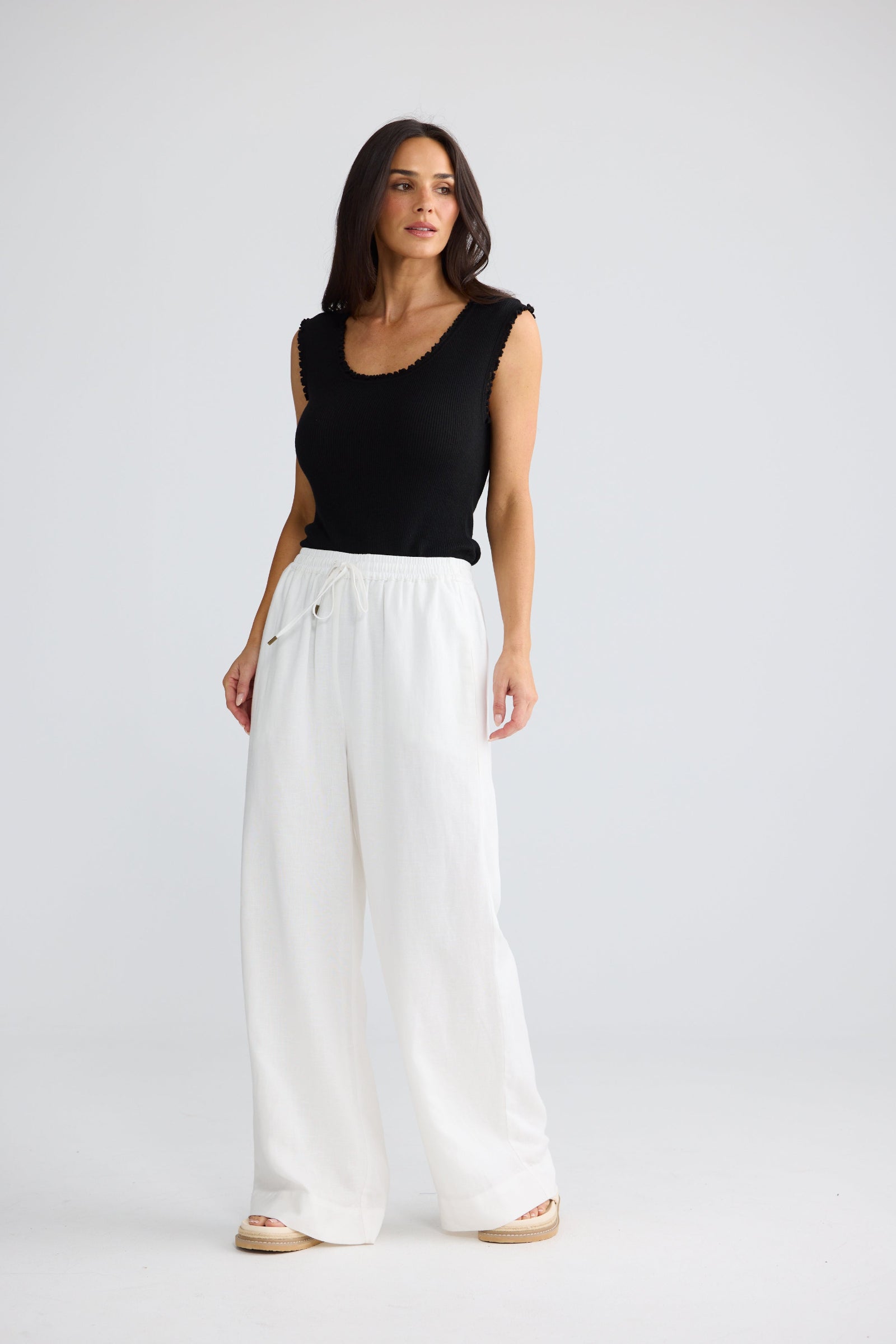 A woman modeling a black sleeveless top and white wide-leg pants against a light gray studio background.