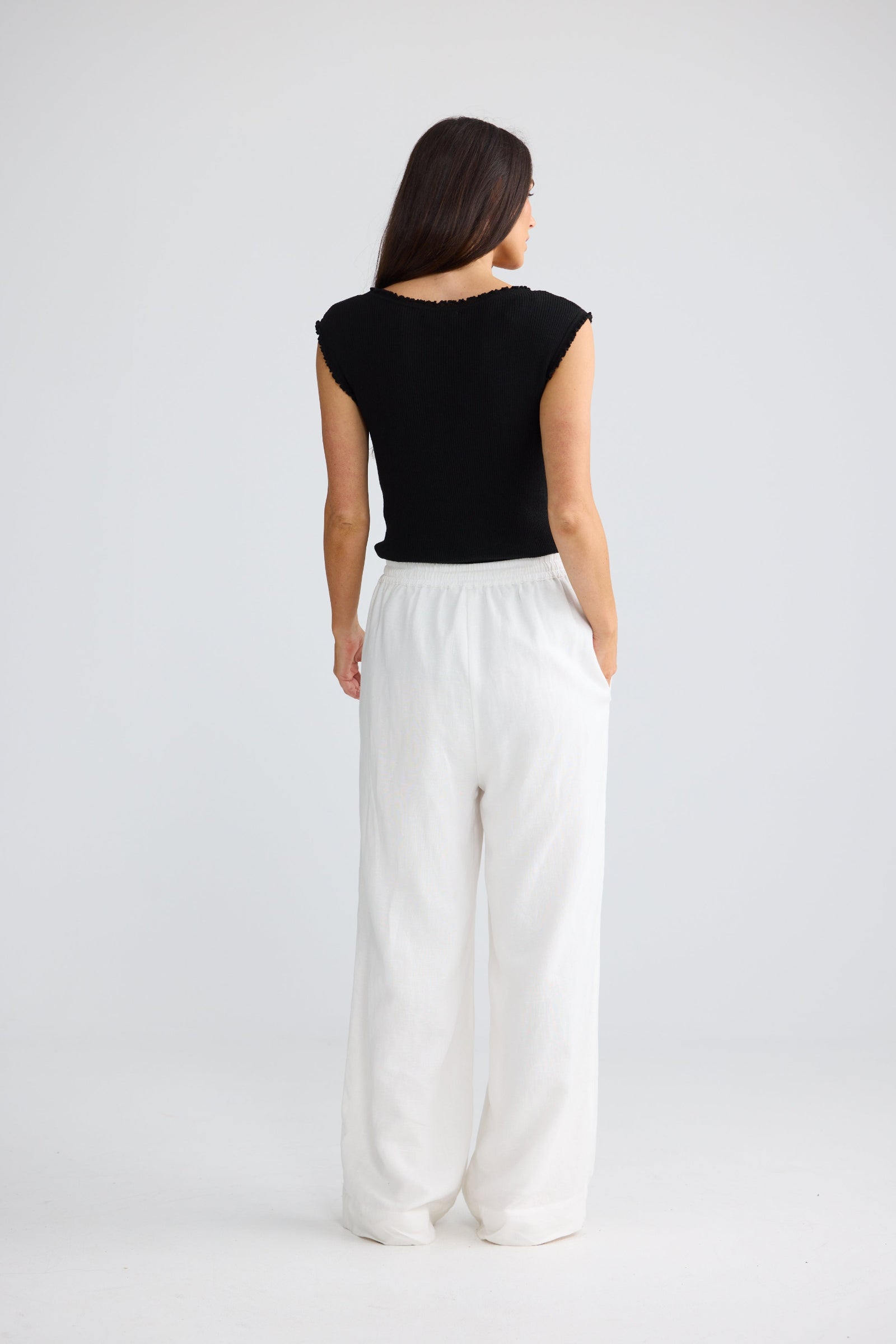 A woman viewed from behind wearing a black sleeveless top and white wide-leg pants against a light gray studio background.