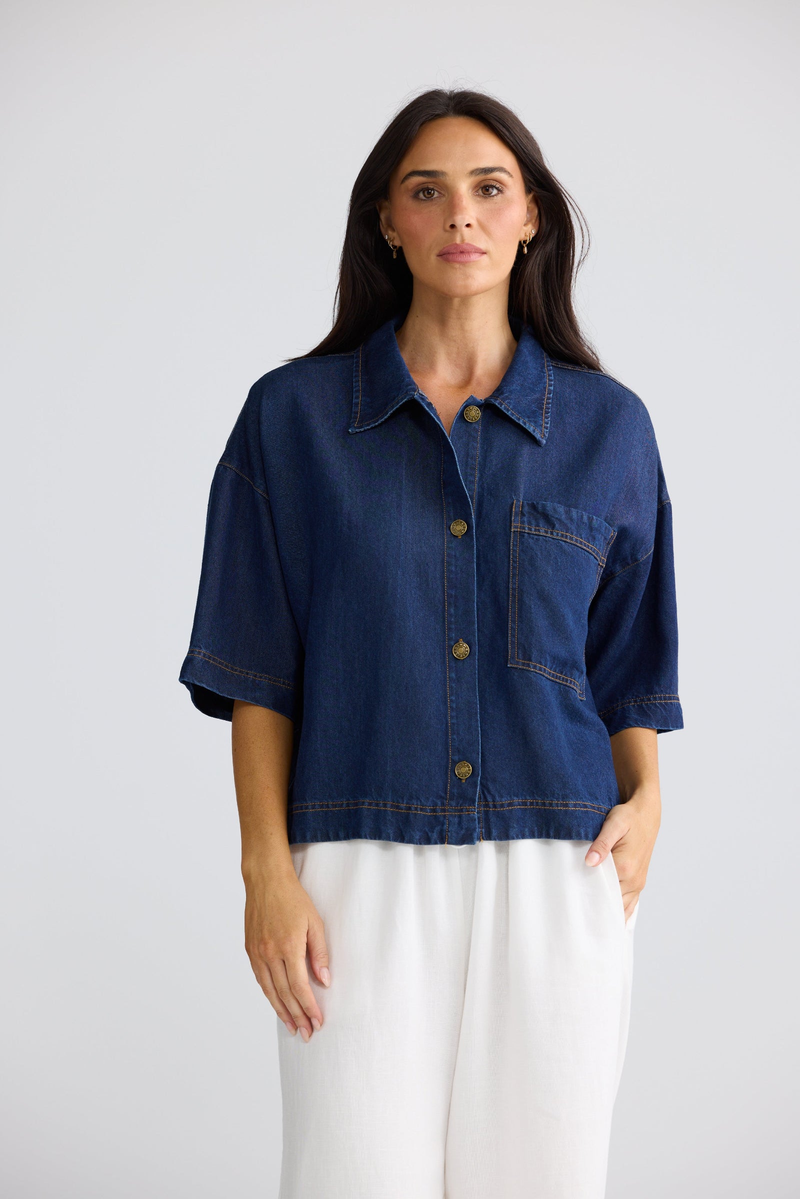 A woman modeling a cropped dark blue denim shirt jacket with three-quarter sleeves, a button-up front, and a chest pocket, paired with white pants, against a light gray studio background.