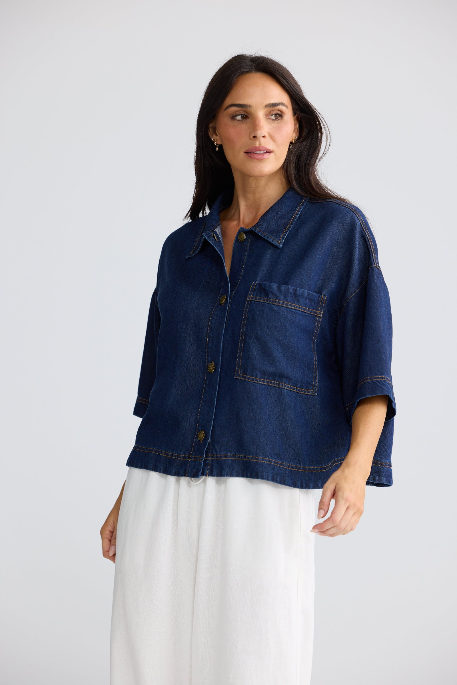 A woman modeling a dark indigo blue cropped denim jacket with three-quarter sleeves and a button-up front, paired with white wide-leg pants, against a light gray studio background.