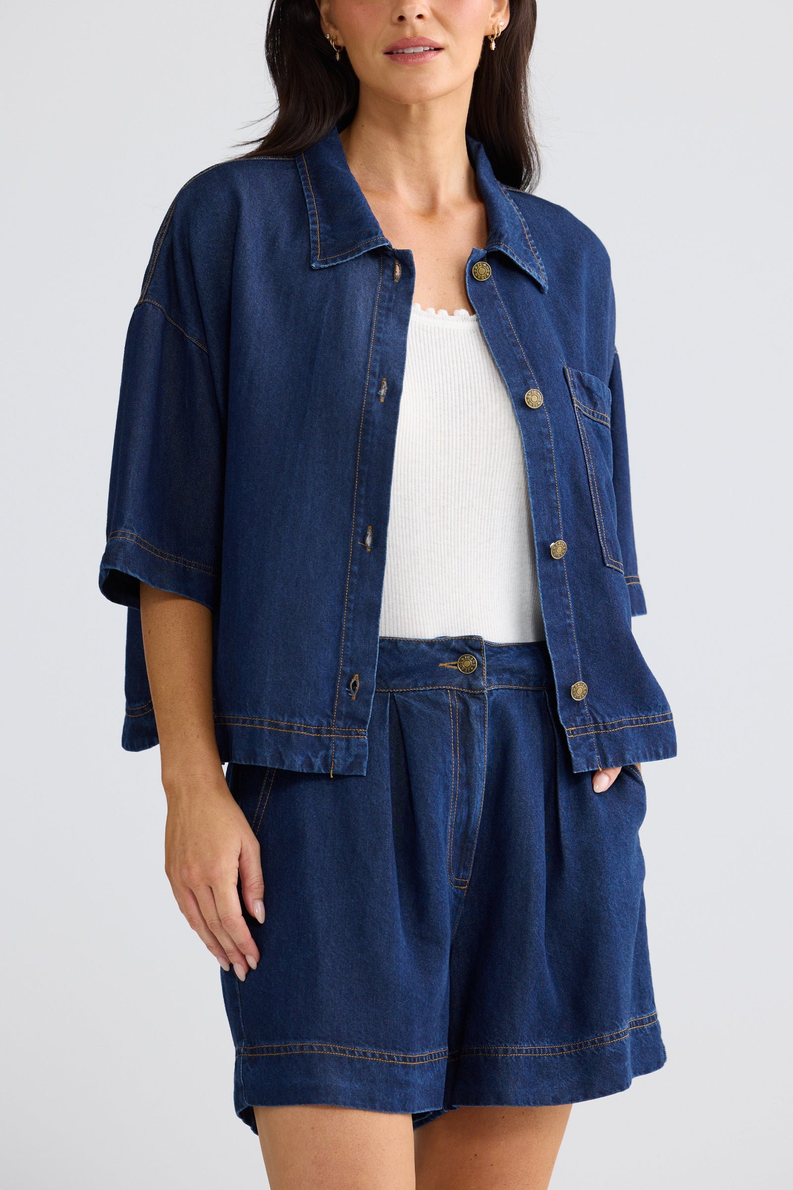 A woman wearing a dark blue denim jacket with three-quarter sleeves and matching denim shorts, styled over a white ribbed top, against a light gray studio background.