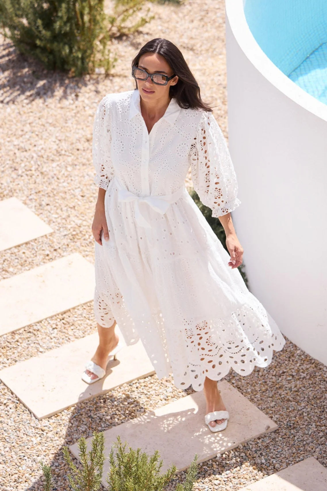 A woman wearing a white eyelet midi dress walks along stone steps beside a white curved wall and blue pool water in a sunny outdoor setting.