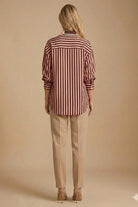 A woman viewed from behind wearing a long-sleeve button-up shirt with vertical burgundy and beige stripes, paired with beige trousers and beige heels, against a solid beige studio background.
