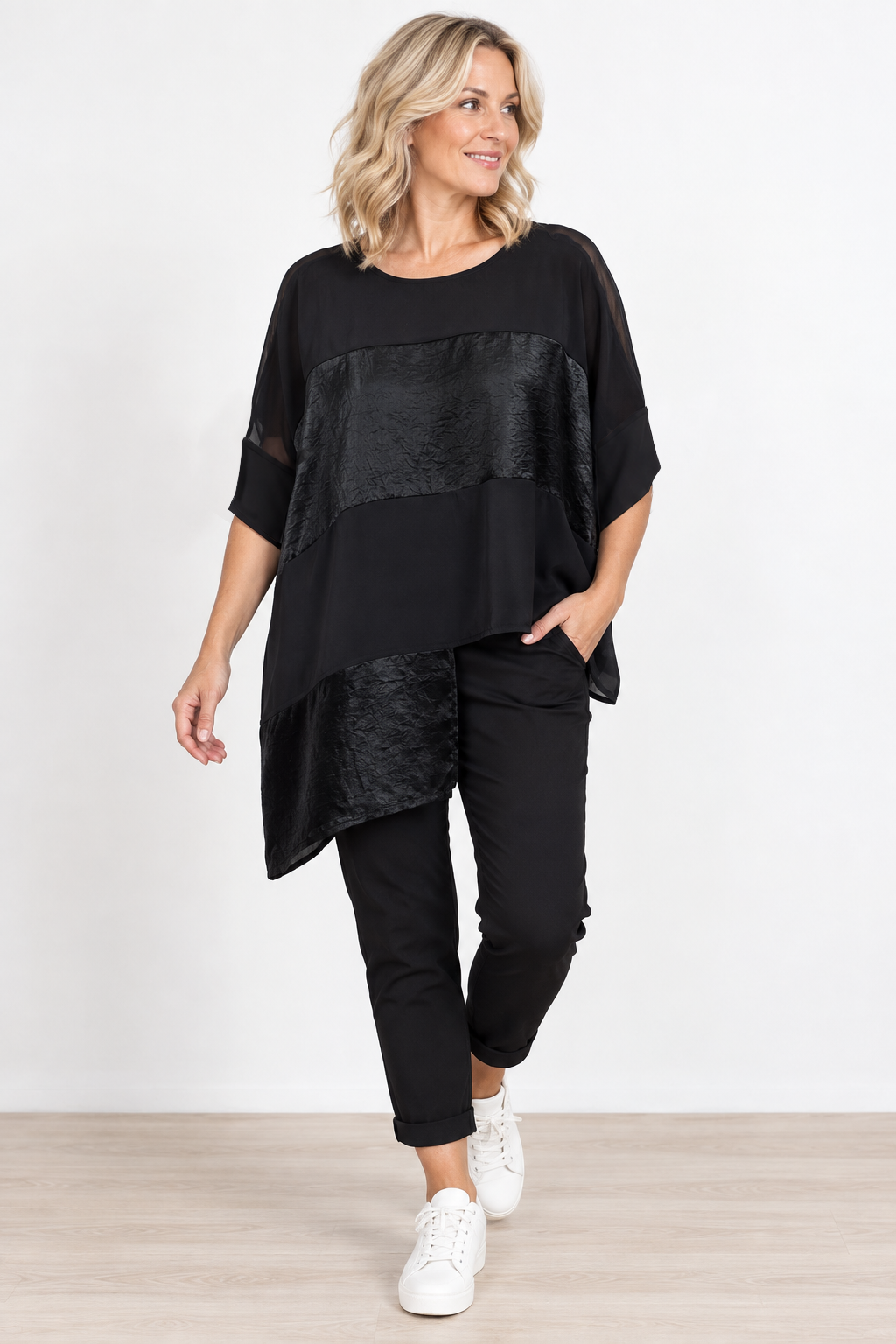 A woman modeling a black asymmetrical short-sleeve top with textured paneling, paired with black pants and white sneakers, against a clean white studio background.